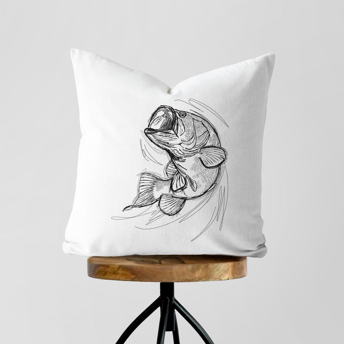 White pillow cover printed with a detailed black ink illustration of a largemouth bass leaping with its mouth open, on a wooden stool.
