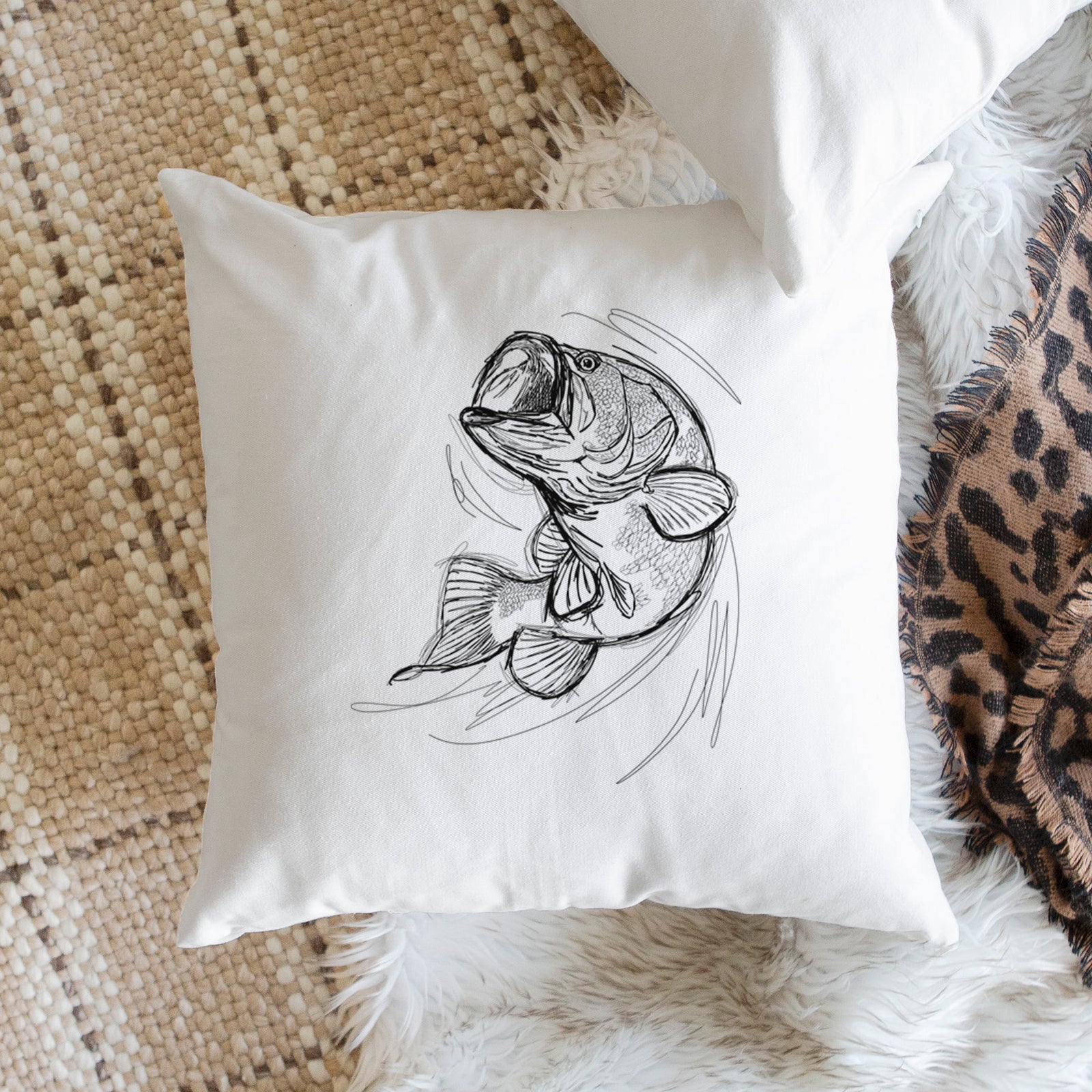 White pillow cover printed with a detailed black ink illustration of a largemouth bass leaping with its mouth open, on a bench beside plants.