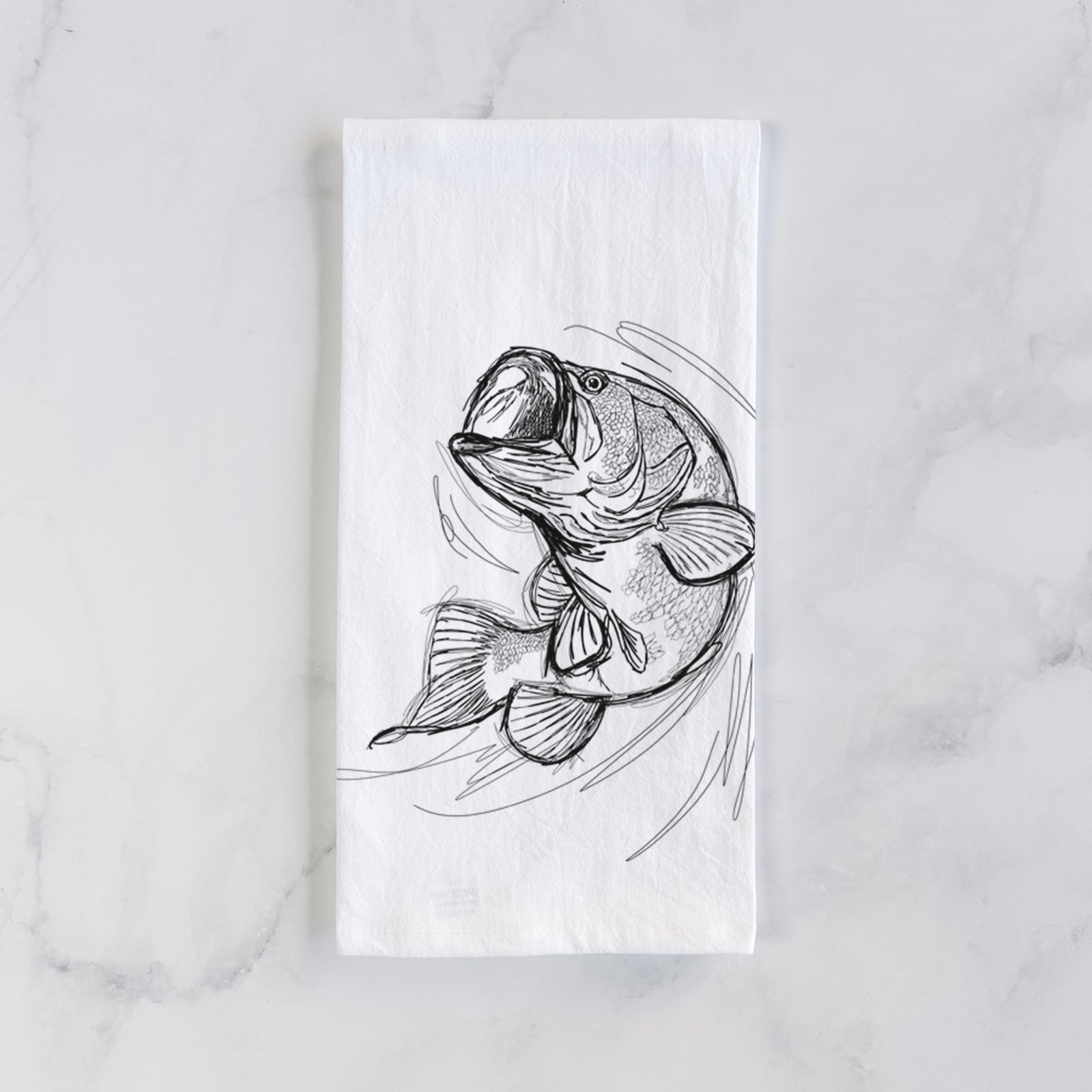 White cotton tea towel printed with a detailed black ink illustration of a largemouth bass leaping with its mouth open, on a marble counter beside a round wooden cutting board and greenery.