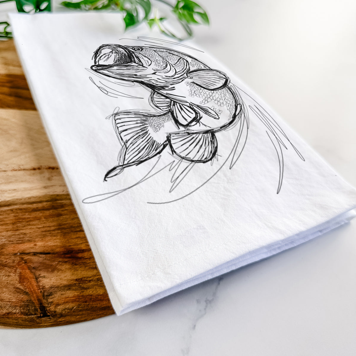 Close-up of a folded white tea towel on a wooden board, showing a detailed black ink illustration of a largemouth bass leaping with its mouth open.