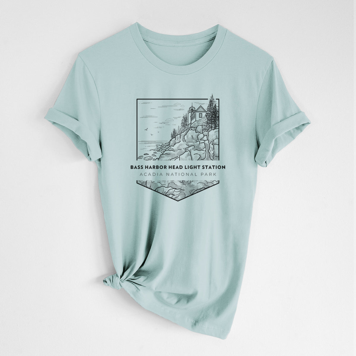 Dusty blue lightweight cotton t-shirt laid flat with the sleeves rolled, printed with a black line art badge of Bass Harbor Head Light Station on a rocky cliff, with the words Acadia National Park.