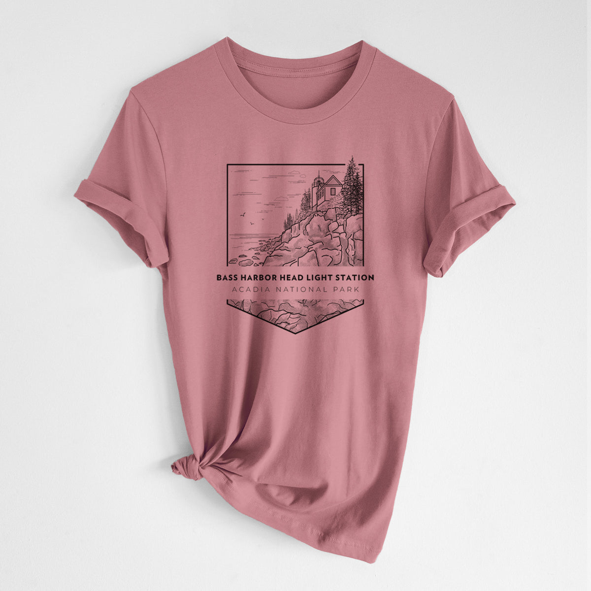 Mauve pink lightweight cotton t-shirt laid flat with the sleeves rolled, printed with a black line art badge of Bass Harbor Head Light Station on a rocky cliff, with the words Acadia National Park.