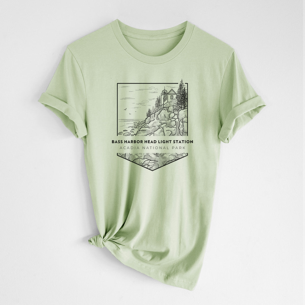 Spring green lightweight cotton t-shirt laid flat with the sleeves rolled, printed with a black line art badge of Bass Harbor Head Light Station on a rocky cliff, with the words Acadia National Park.