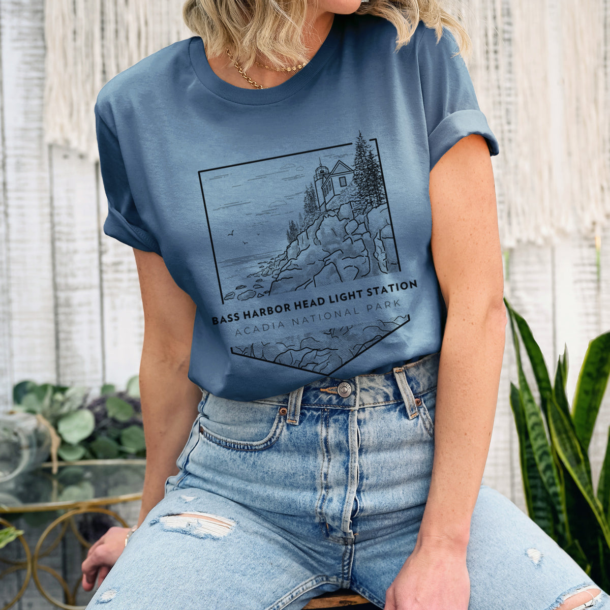 Woman wearing a steel blue lightweight cotton t-shirt printed with a black line art badge of Bass Harbor Head Light Station on a rocky cliff, with the words Acadia National Park.