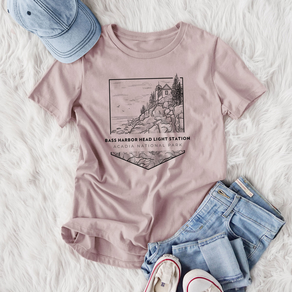 Women's relaxed fit cotton t-shirt printed with a black line art badge of Bass Harbor Head Light Station on a rocky cliff, with the words Acadia National Park, styled flat on a white faux fur rug with jeans.