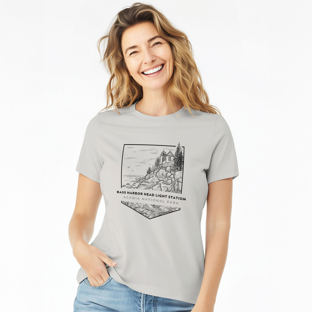 Woman wearing a lunar grey women's relaxed fit cotton t-shirt printed with a black line art badge of Bass Harbor Head Light Station on a rocky cliff, with the words Acadia National Park.