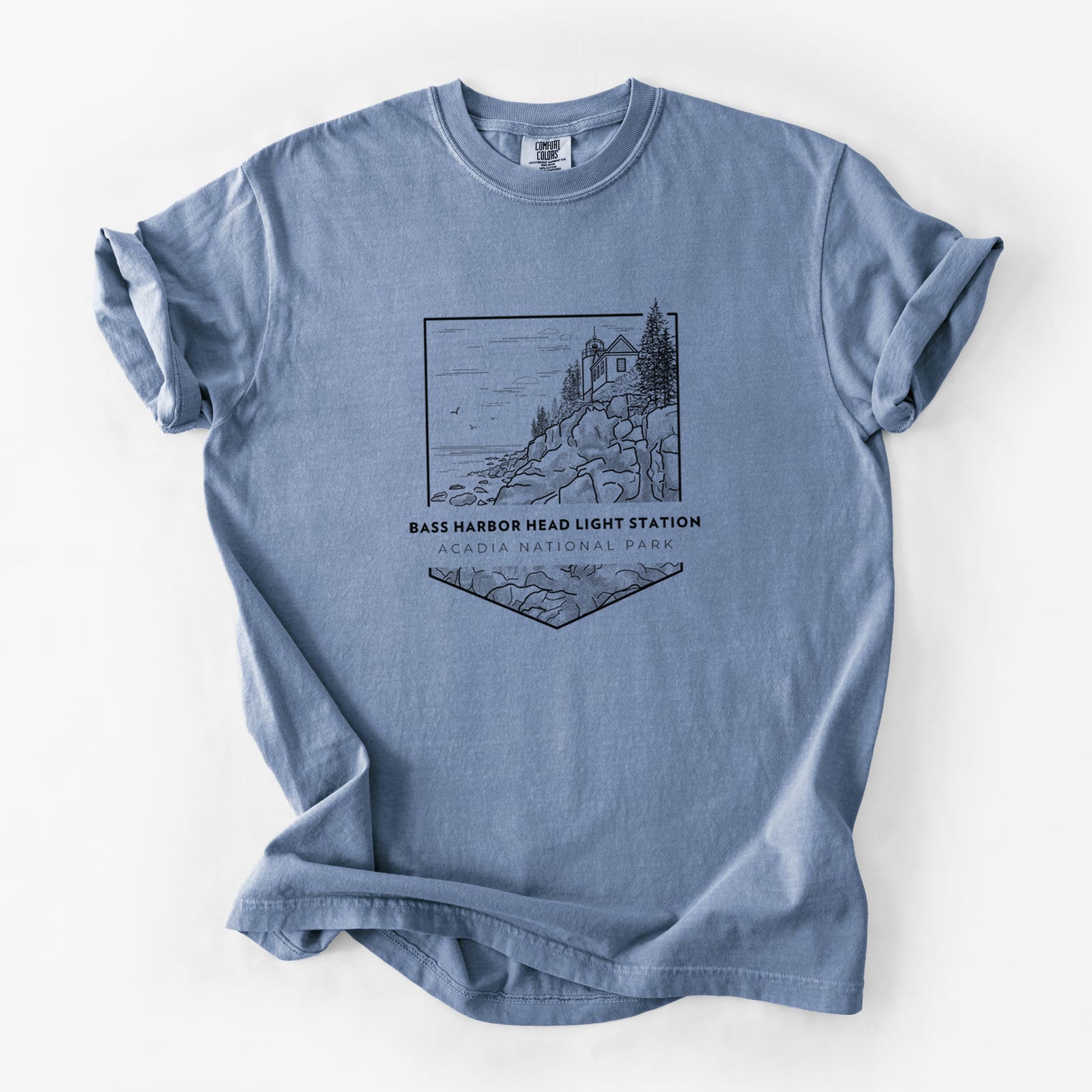 Blue jean Comfort Colors t-shirt laid flat, printed with a black line art badge of Bass Harbor Head Light Station on a rocky cliff, with the words Acadia National Park.