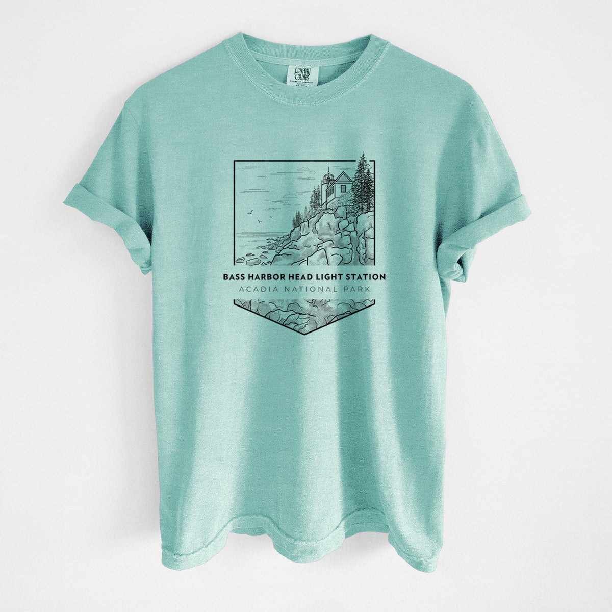 Chalky mint Comfort Colors t-shirt laid flat, printed with a black line art badge of Bass Harbor Head Light Station on a rocky cliff, with the words Acadia National Park.