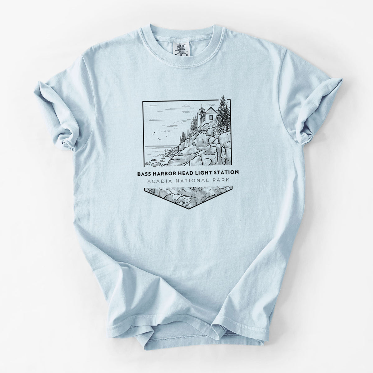 Chambray blue Comfort Colors t-shirt laid flat, printed with a black line art badge of Bass Harbor Head Light Station on a rocky cliff, with the words Acadia National Park.