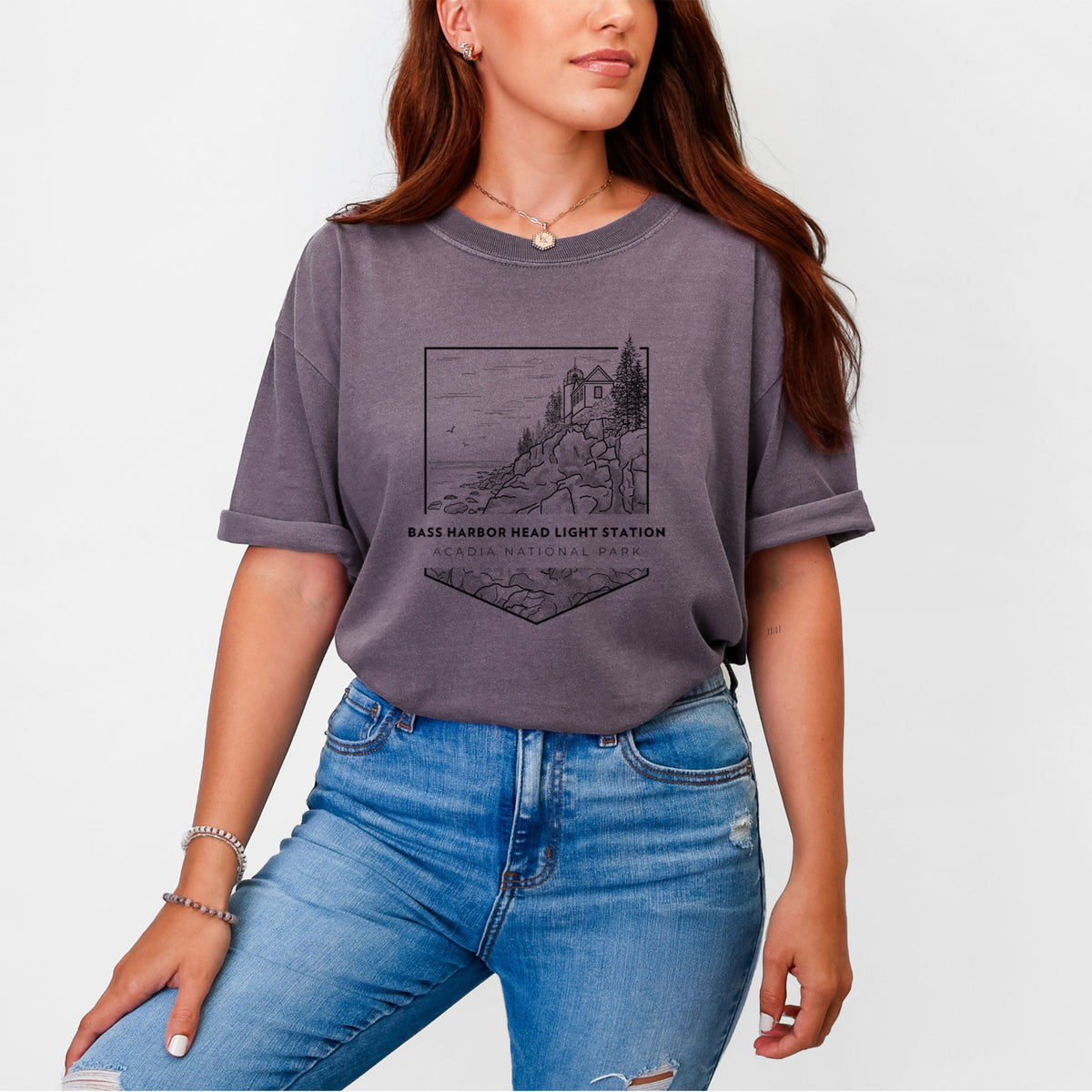 Woman wearing a Comfort Colors t-shirt printed with a black line art badge of Bass Harbor Head Light Station on a rocky cliff, with the words Acadia National Park.