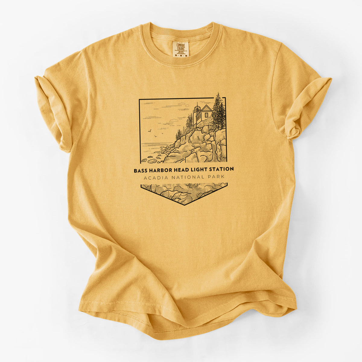 Golden wheat yellow Comfort Colors t-shirt laid flat, printed with a black line art badge of Bass Harbor Head Light Station on a rocky cliff, with the words Acadia National Park.