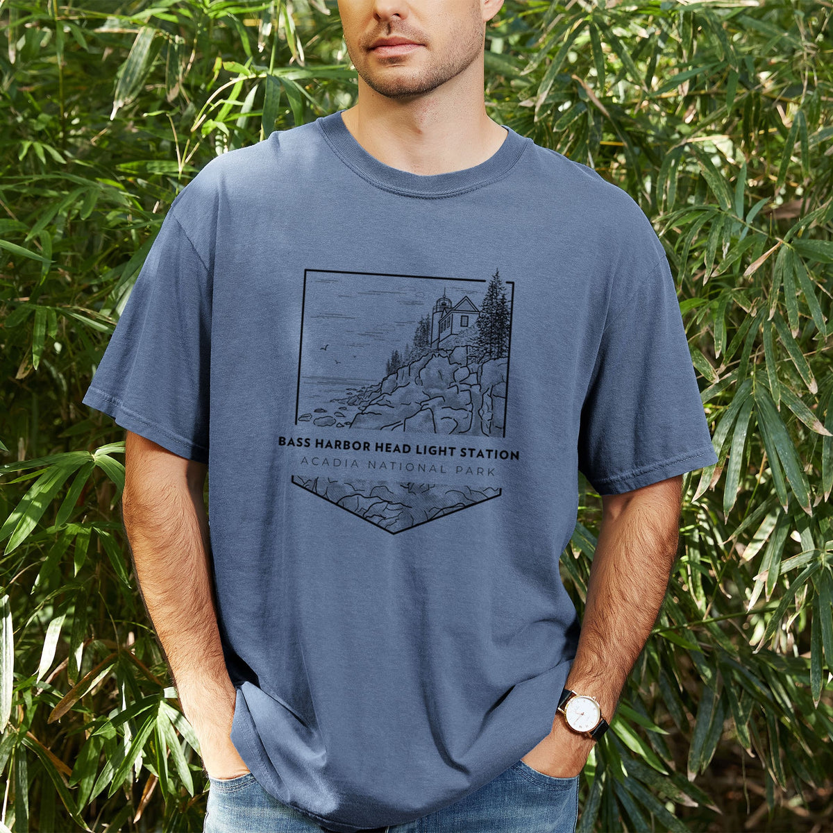 Man wearing a Comfort Colors t-shirt printed with a black line art badge of Bass Harbor Head Light Station on a rocky cliff, with the words Acadia National Park, outdoors.