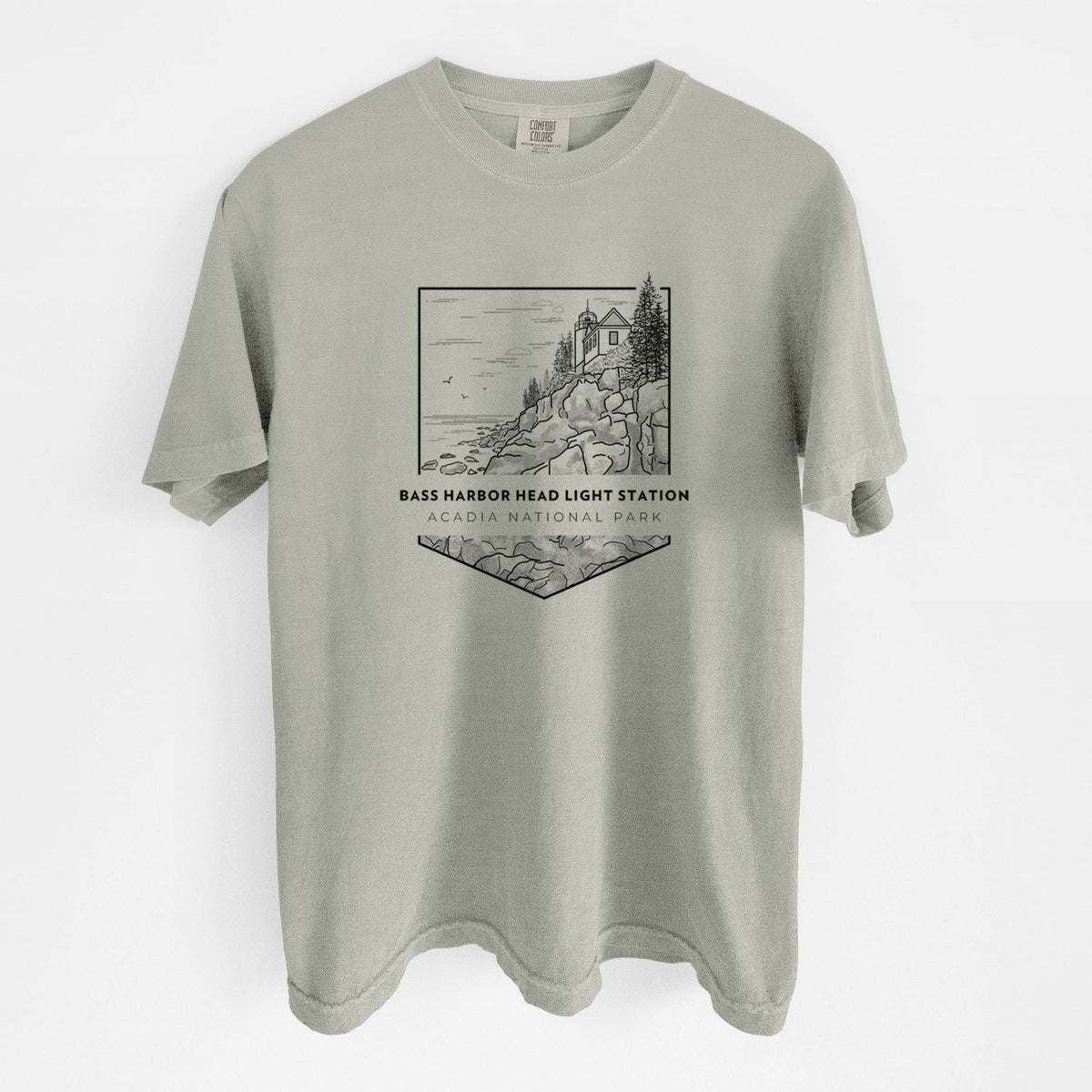 Sandstone beige Comfort Colors t-shirt laid flat, printed with a black line art badge of Bass Harbor Head Light Station on a rocky cliff, with the words Acadia National Park.