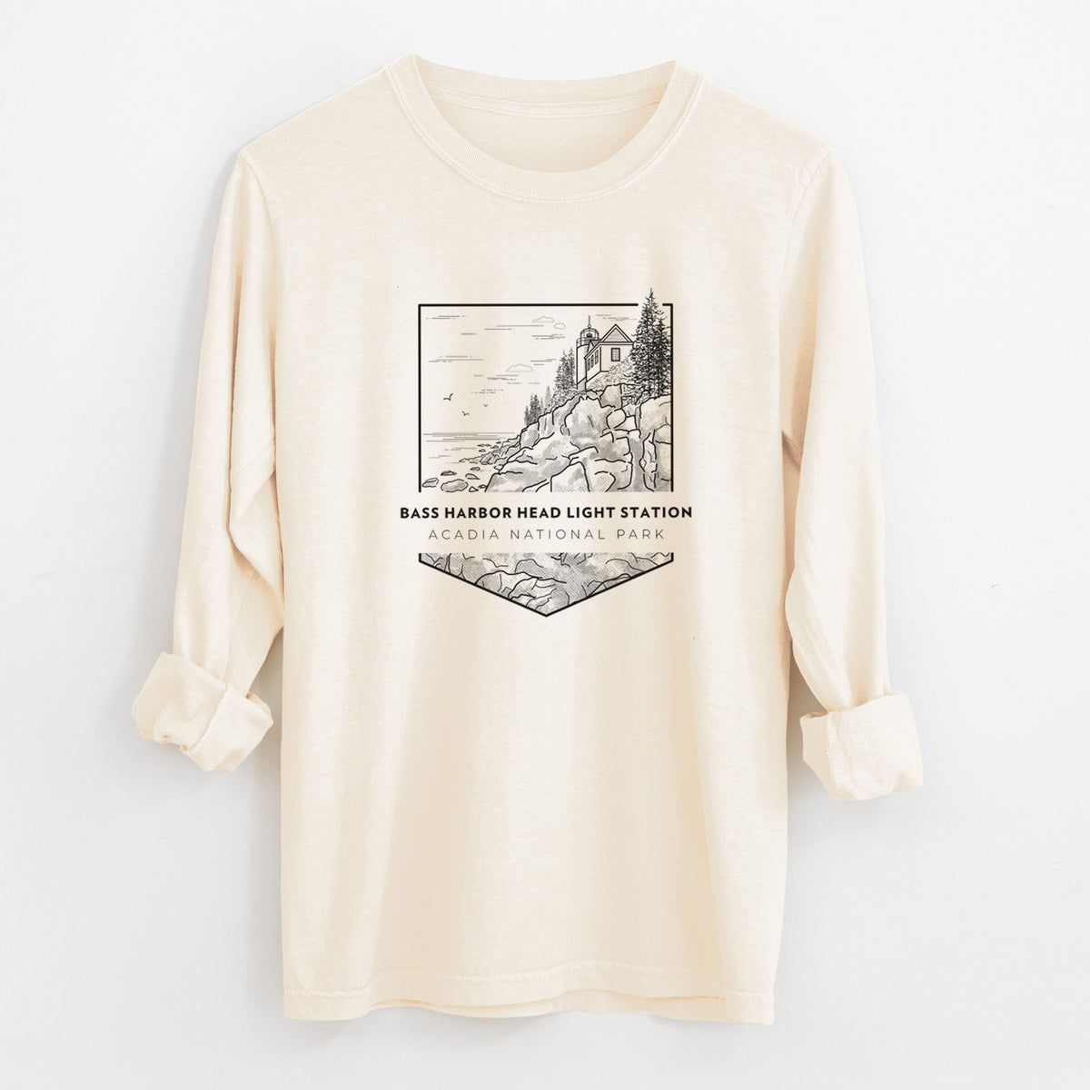 Ivory Comfort Colors long sleeve shirt laid flat with the cuffs rolled, printed with a black line art badge of Bass Harbor Head Light Station on a rocky cliff, with the words Acadia National Park.