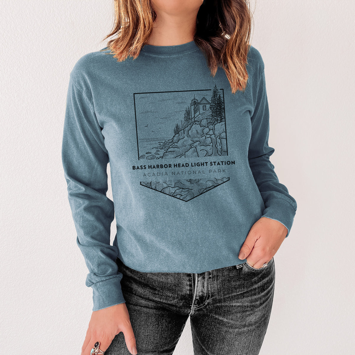 Steel blue Comfort Colors long sleeve shirt printed with a black line art badge of Bass Harbor Head Light Station on a rocky cliff, with the words Acadia National Park, shown in a lifestyle photo.