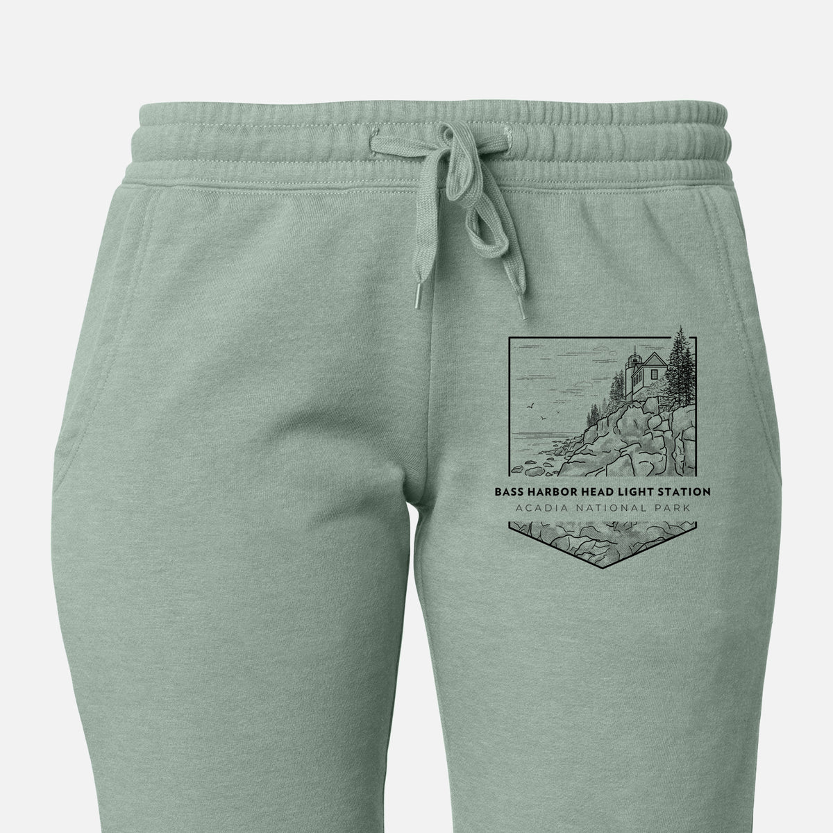 Front of sage green Cali Wave sweatpants, printed with a black line art badge of Bass Harbor Head Light Station on a rocky cliff, with the words Acadia National Park on the leg.