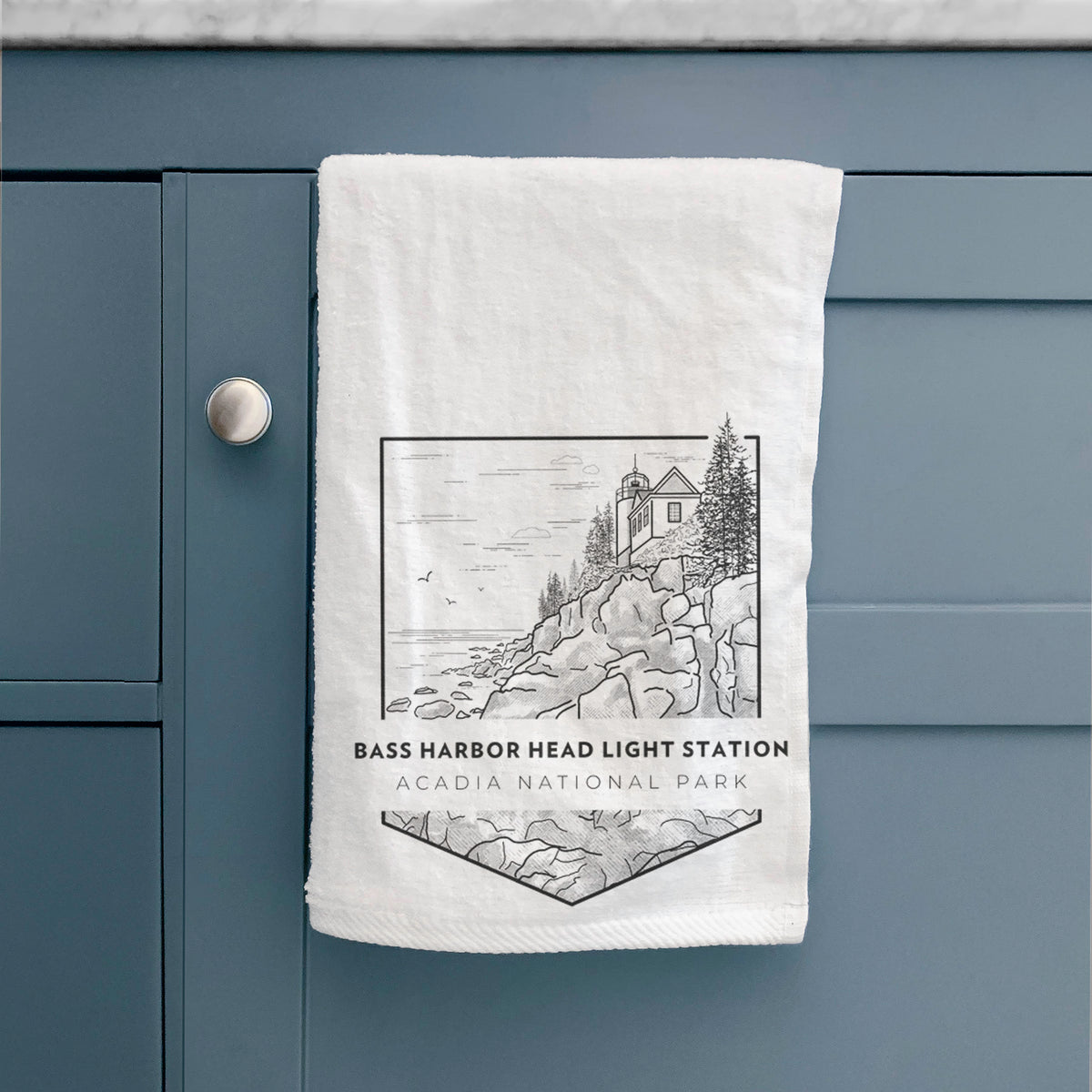 White hand towel printed with a black line art badge of Bass Harbor Head Light Station on a rocky cliff, with the words Acadia National Park, hanging on a cabinet door.
