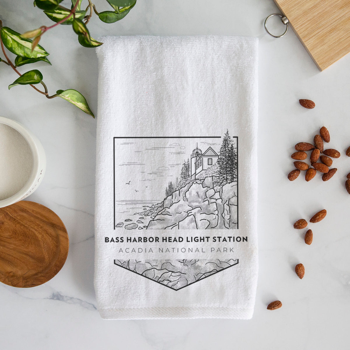 White hand towel printed with a black line art badge of Bass Harbor Head Light Station on a rocky cliff, with the words Acadia National Park.