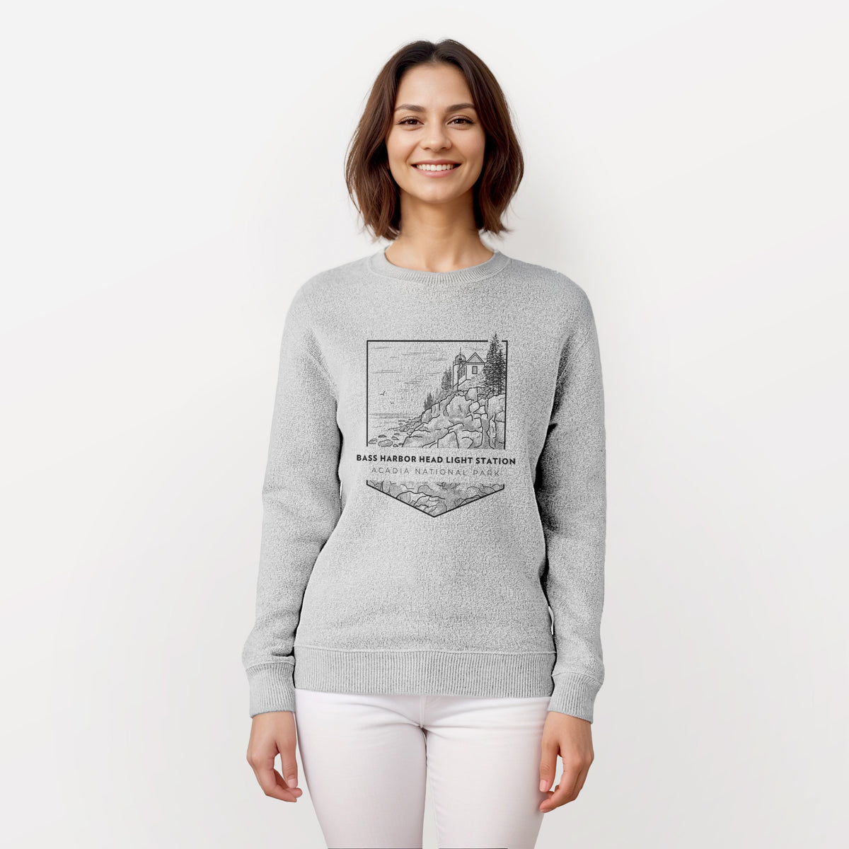 Woman wearing a knit sweatshirt printed with a black line art badge of Bass Harbor Head Light Station on a rocky cliff, with the words Acadia National Park.