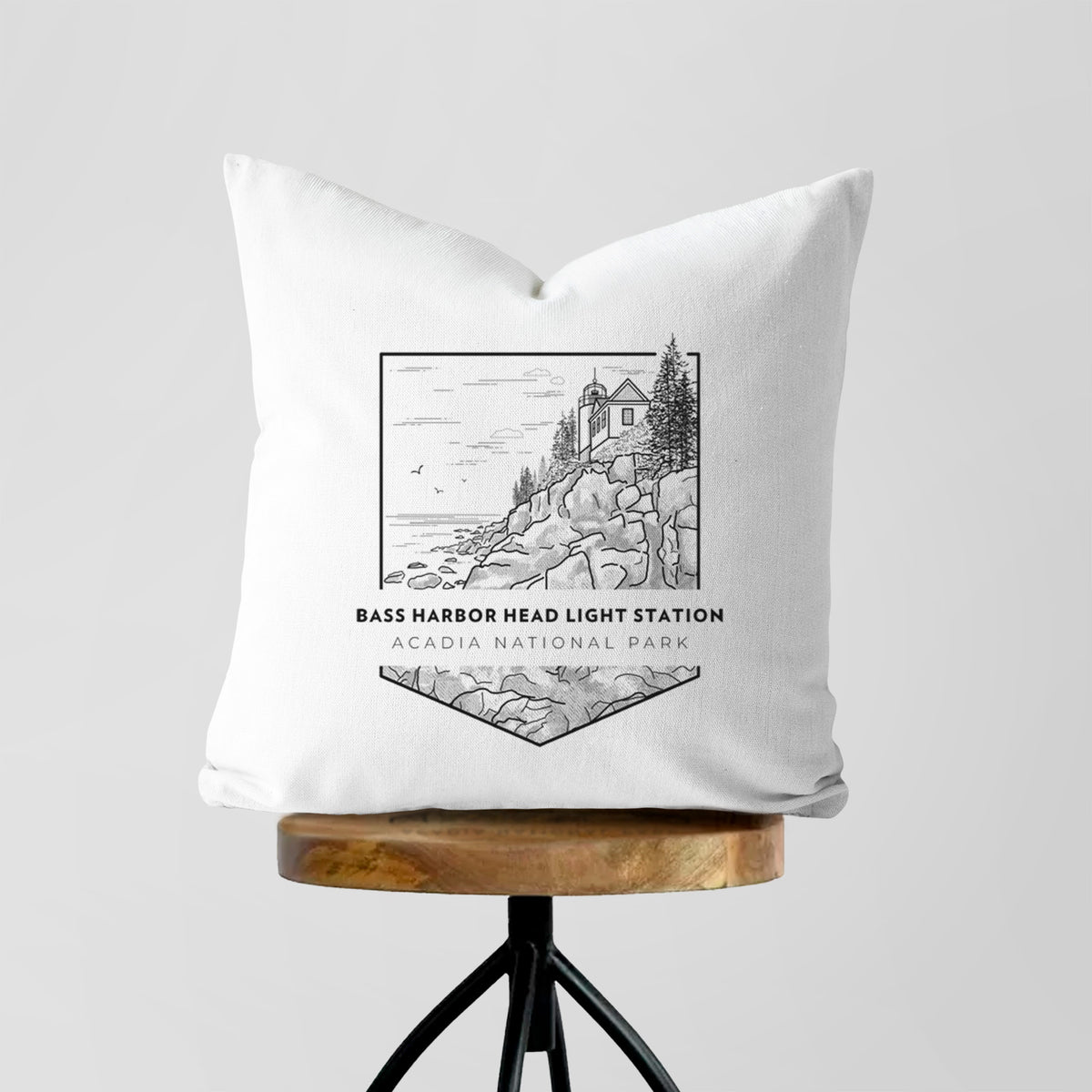 White pillow cover printed with a black line art badge of Bass Harbor Head Light Station on a rocky cliff, with the words Acadia National Park, on a wooden stool.