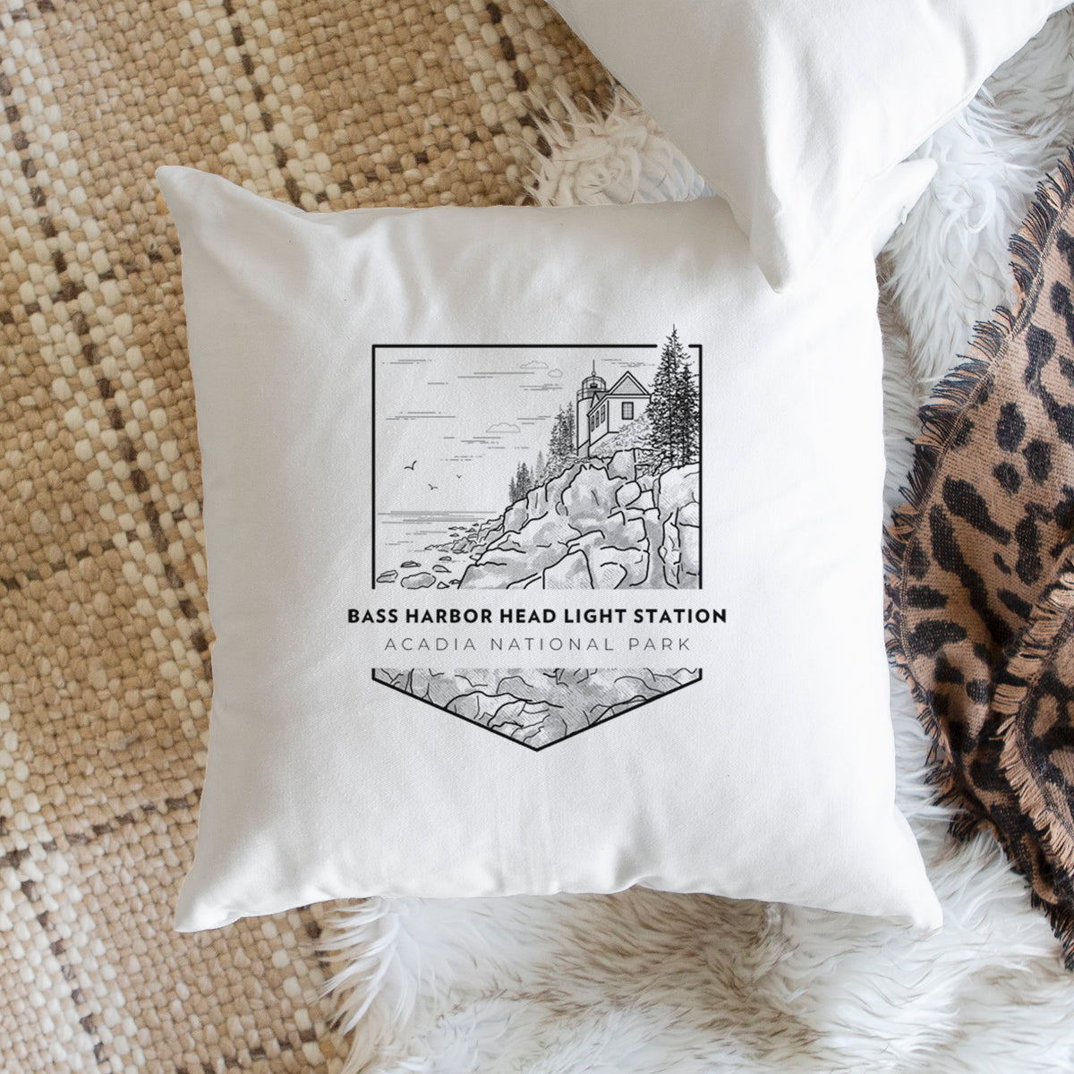 White pillow cover printed with a black line art badge of Bass Harbor Head Light Station on a rocky cliff, with the words Acadia National Park, on a woven chair.
