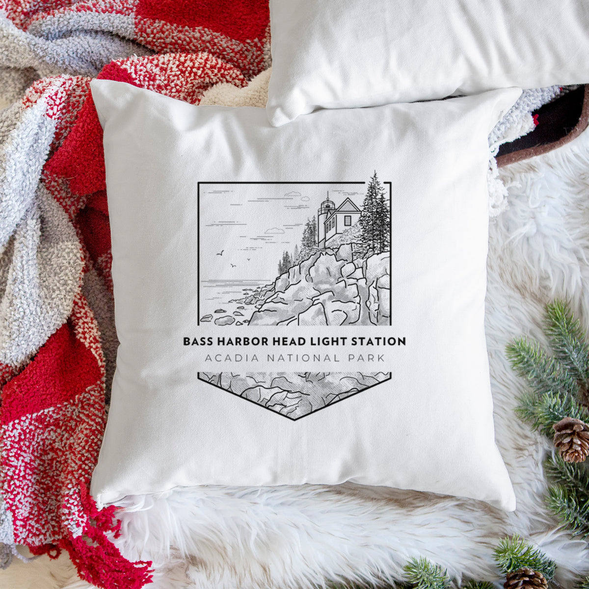 White pillow cover printed with a black line art badge of Bass Harbor Head Light Station on a rocky cliff, with the words Acadia National Park, on a woven chair.