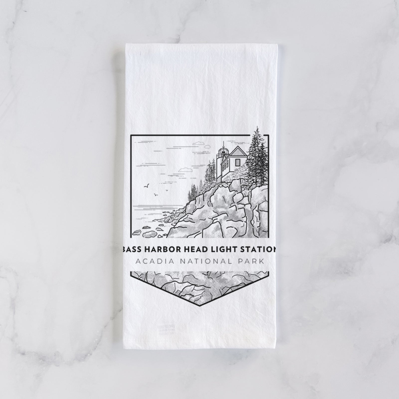 White cotton tea towel printed with a black line art badge of Bass Harbor Head Light Station on a rocky cliff, with the words Acadia National Park, on a marble counter beside a round wooden cutting board and greenery.