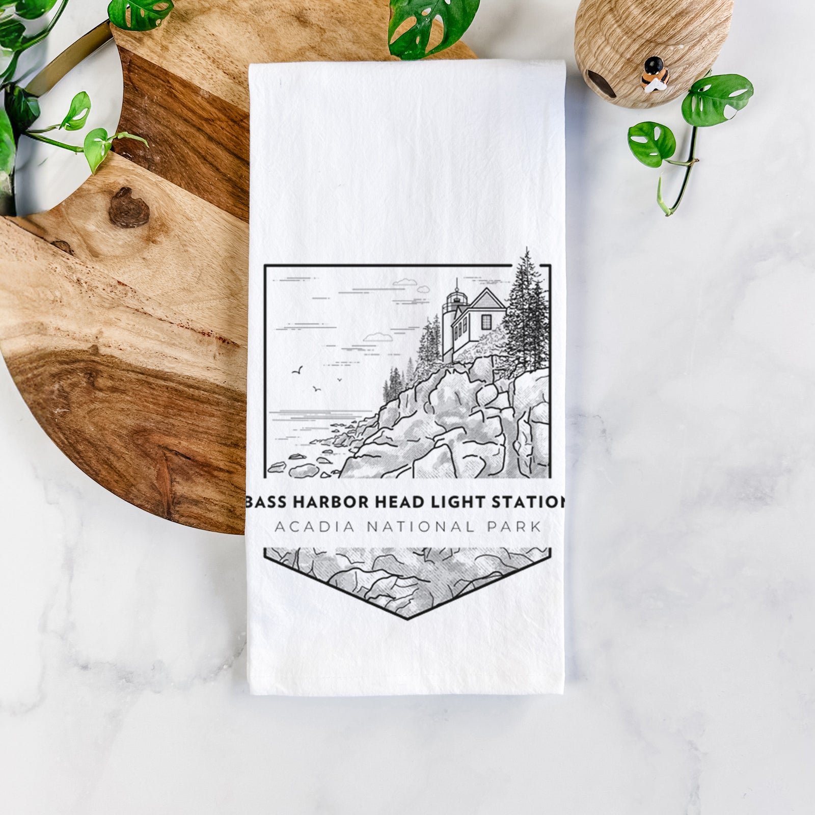 White cotton tea towel printed with a black line art badge of Bass Harbor Head Light Station on a rocky cliff, with the words Acadia National Park, on a marble counter beside a round wooden cutting board and greenery.