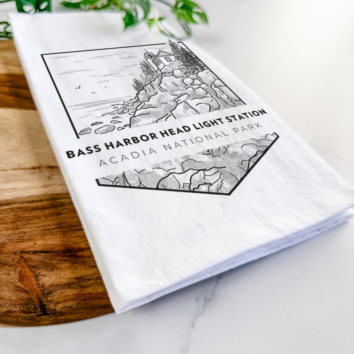 Close-up of a folded white tea towel on a wooden board, showing a black line art badge of Bass Harbor Head Light Station on a rocky cliff, with the words Acadia National Park.