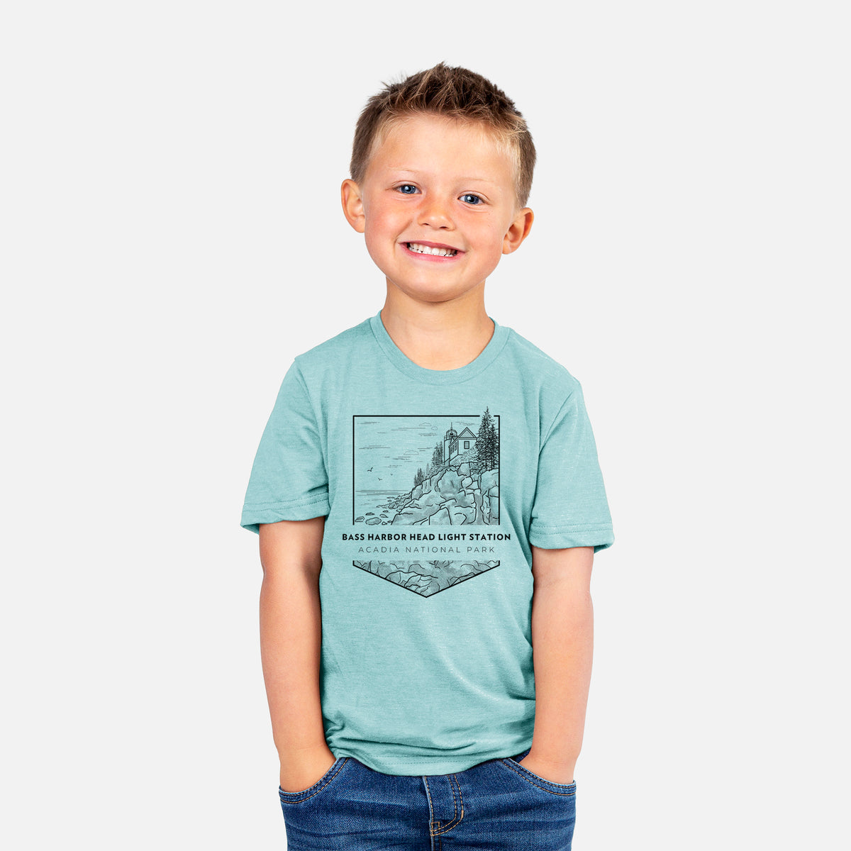 Smiling boy wearing a youth t-shirt printed with a black line art badge of Bass Harbor Head Light Station on a rocky cliff, with the words Acadia National Park.