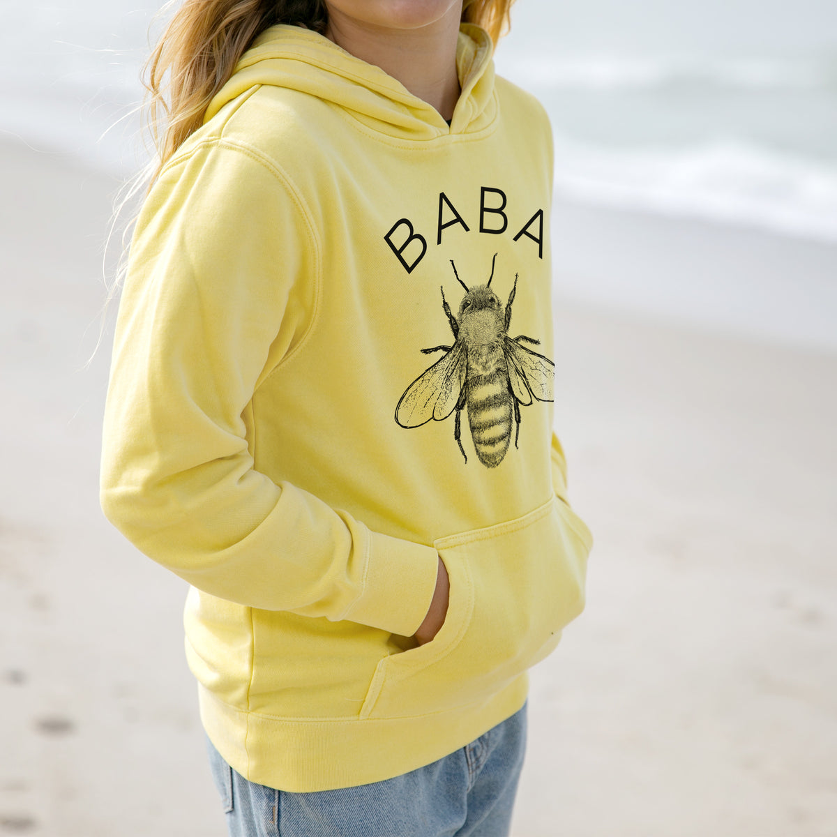 Girl wearing a youth pigment dyed hoodie printed with a black ink honey bee beneath the word Baba.
