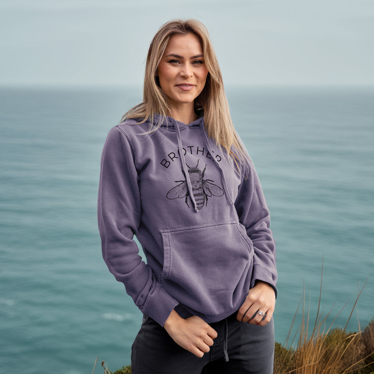 Woman wearing a plum purple pigment dyed hoodie printed with a black ink honey bee beneath the word Brother.