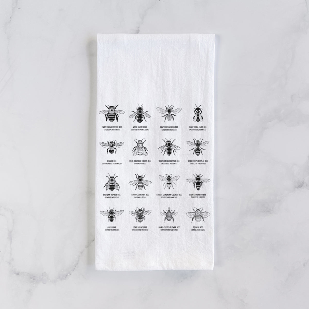 White cotton tea towel laid flat on marble, printed with a chart of black ink illustrations of different bee species with their names.