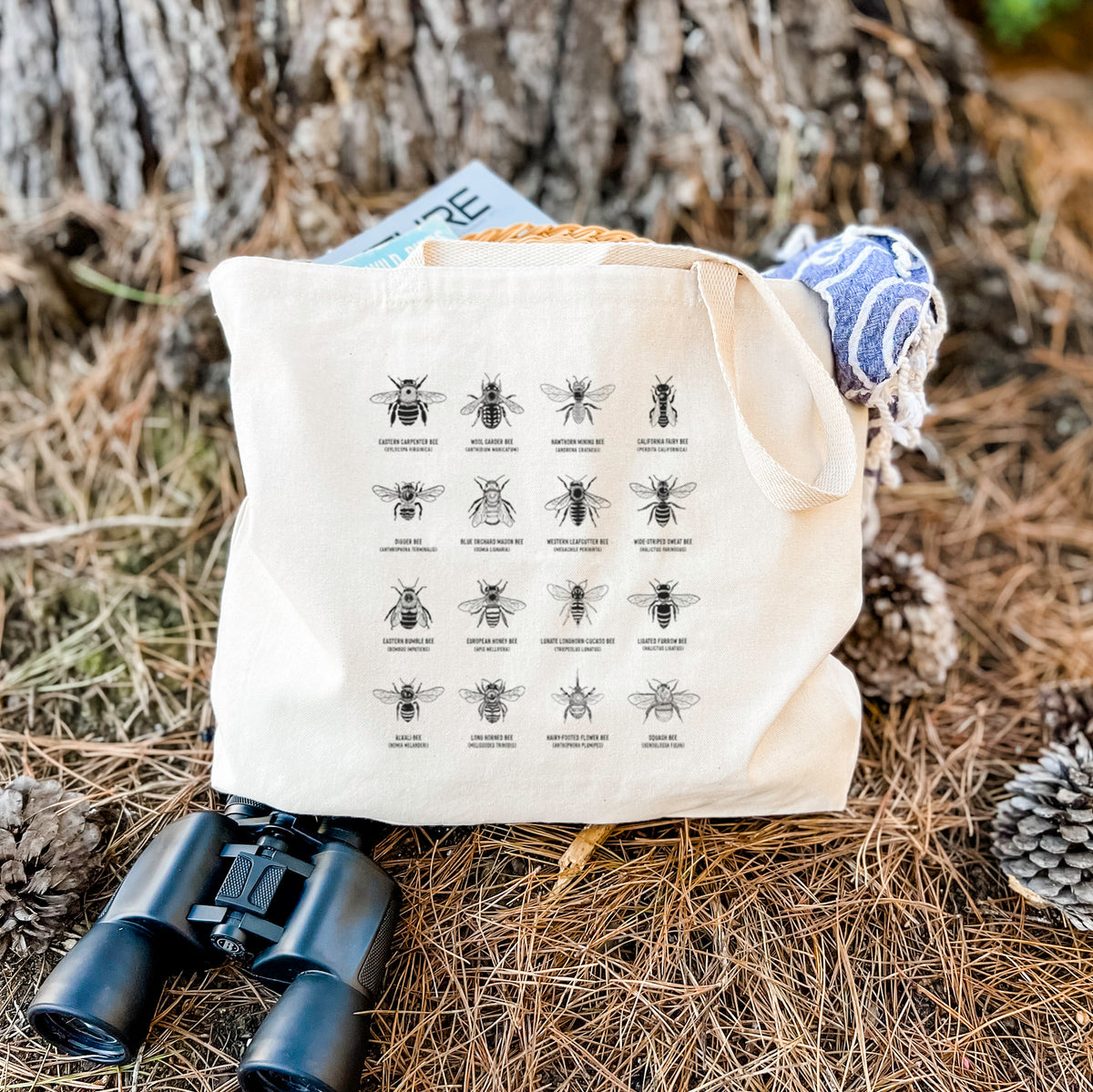 Canvas tote bag printed with a chart of black ink illustrations of different bee species with their names, resting on dry grass beside a pair of binoculars.