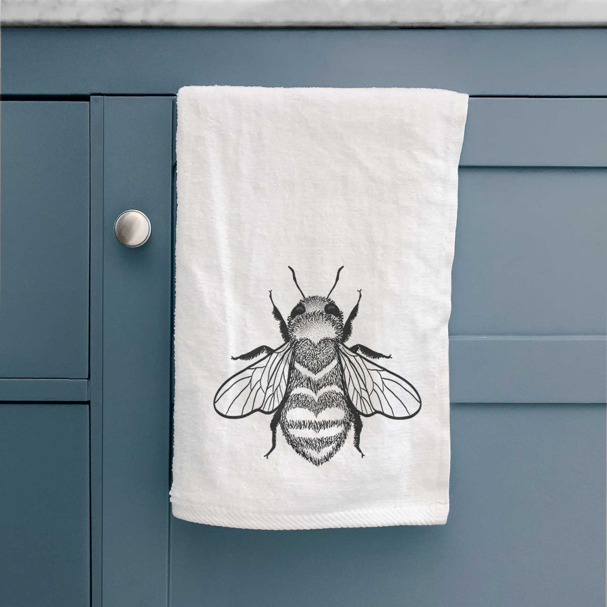 White hand towel printed with a black ink bee with a heart pattern on its body, hanging on a cabinet door.