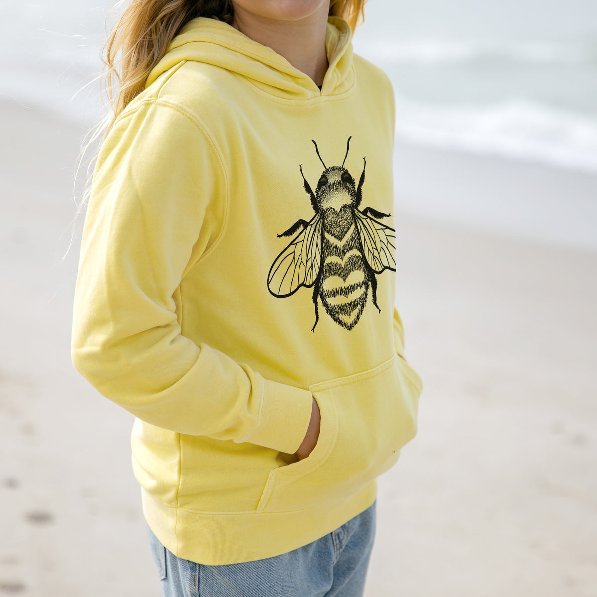 Girl wearing a youth pigment dyed hoodie printed with a black ink bee with a heart pattern on its body.