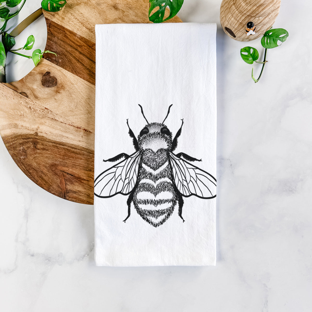 White cotton tea towel printed with a black ink bee with a heart pattern on its body, on a marble counter beside a round wooden cutting board and greenery.