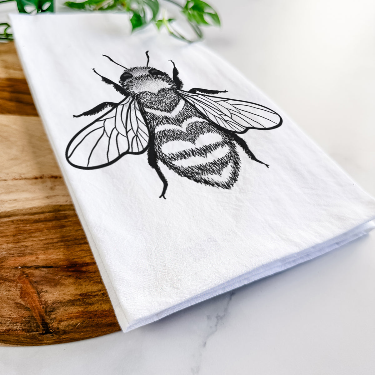 Close-up of a folded white tea towel on a wooden board, showing a black ink bee with a heart pattern on its body.