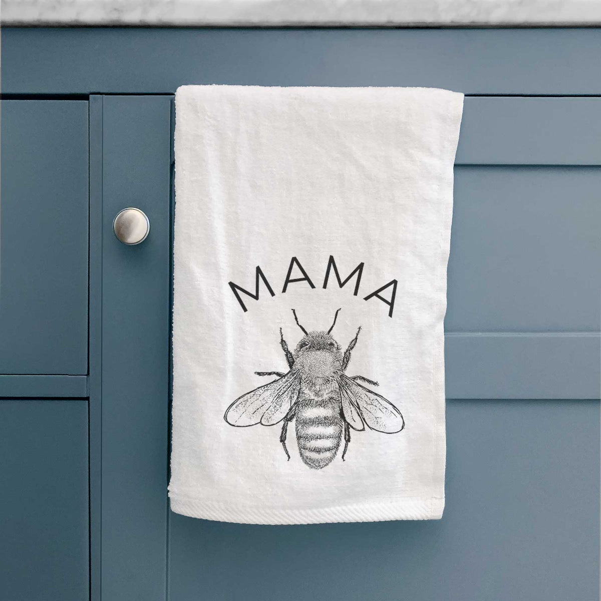 White hand towel printed with a black ink honey bee beneath the word Mama, hanging on a cabinet door.