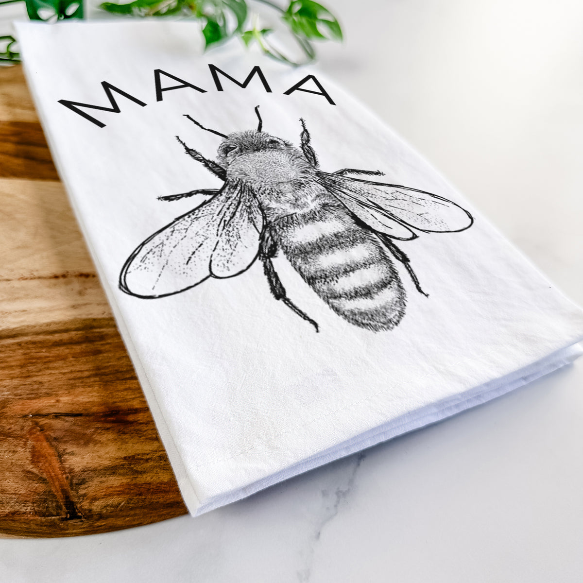 Close-up of a folded white tea towel on a wooden board, showing a black ink honey bee beneath the word Mama.