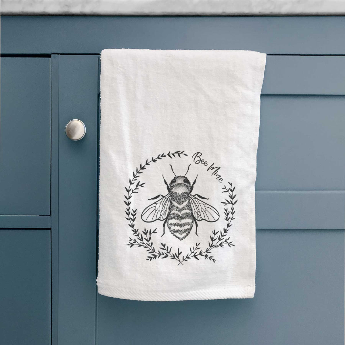 White hand towel printed with a black ink bee inside a wreath of leaves with the words Bee Mine, hanging on a cabinet door.