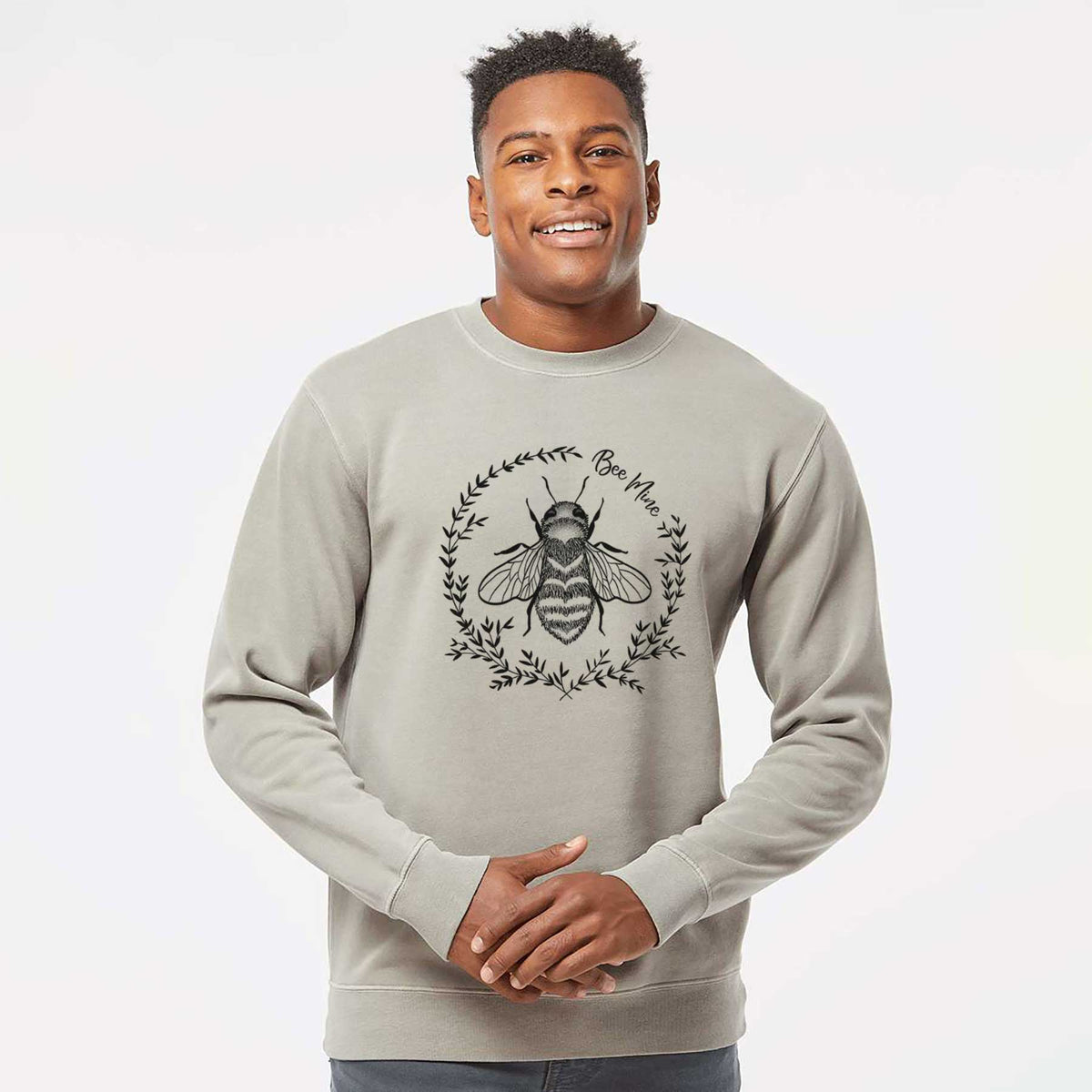 Model wearing a cement grey pigment dyed crewneck sweatshirt printed with a black ink bee inside a wreath of leaves with the words Bee Mine.
