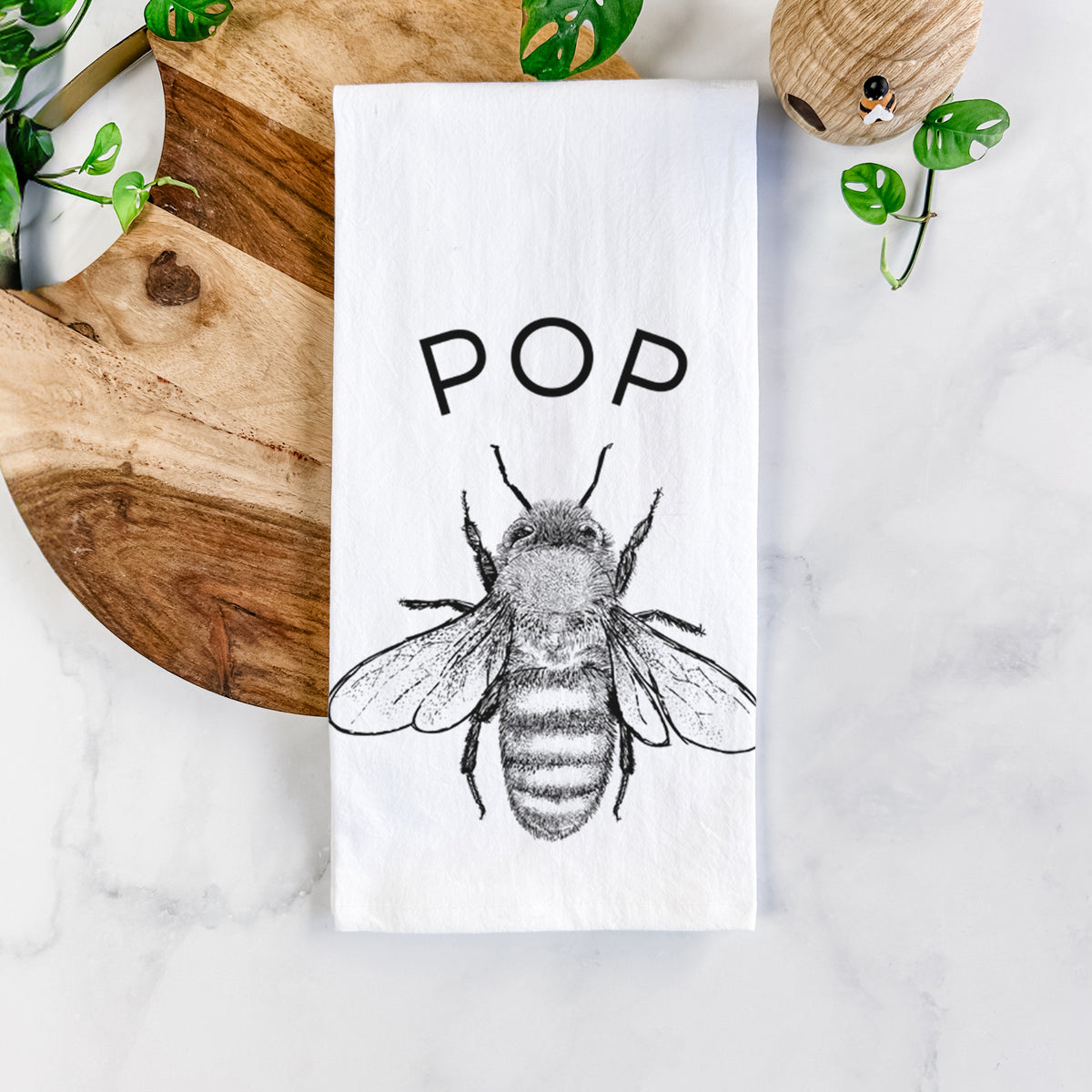White cotton tea towel printed with a black ink honey bee beneath the word Pop, on a marble counter beside a round wooden cutting board and greenery.