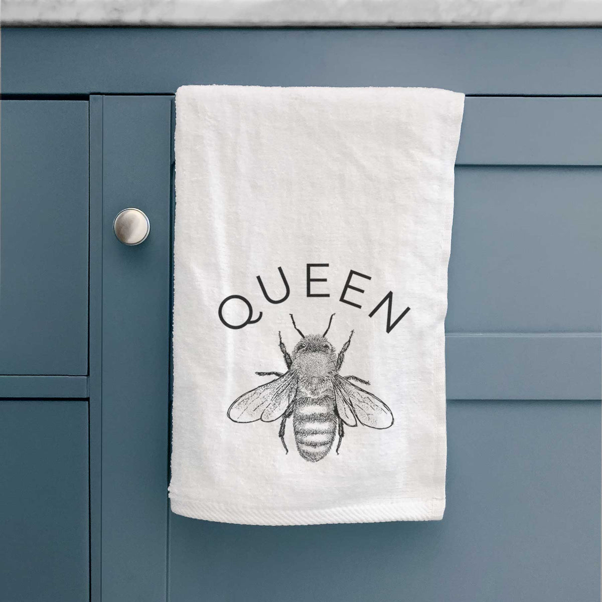 White hand towel printed with a black ink honey bee beneath the word Queen, hanging on a cabinet door.