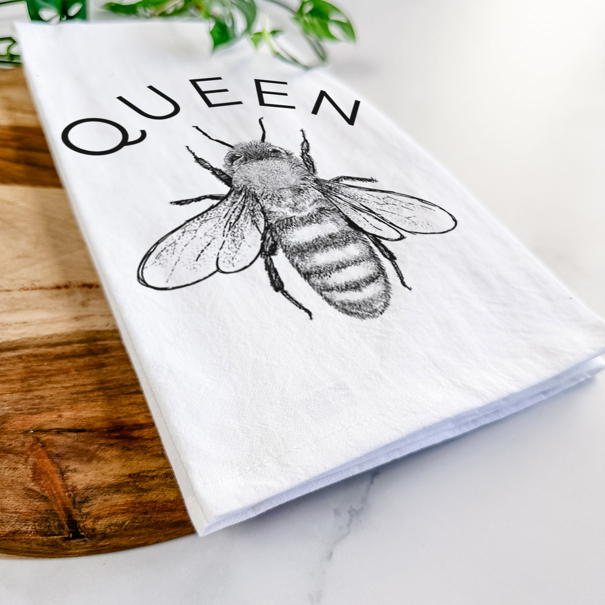 Close-up of a folded white tea towel on a wooden board, showing a black ink honey bee beneath the word Queen.