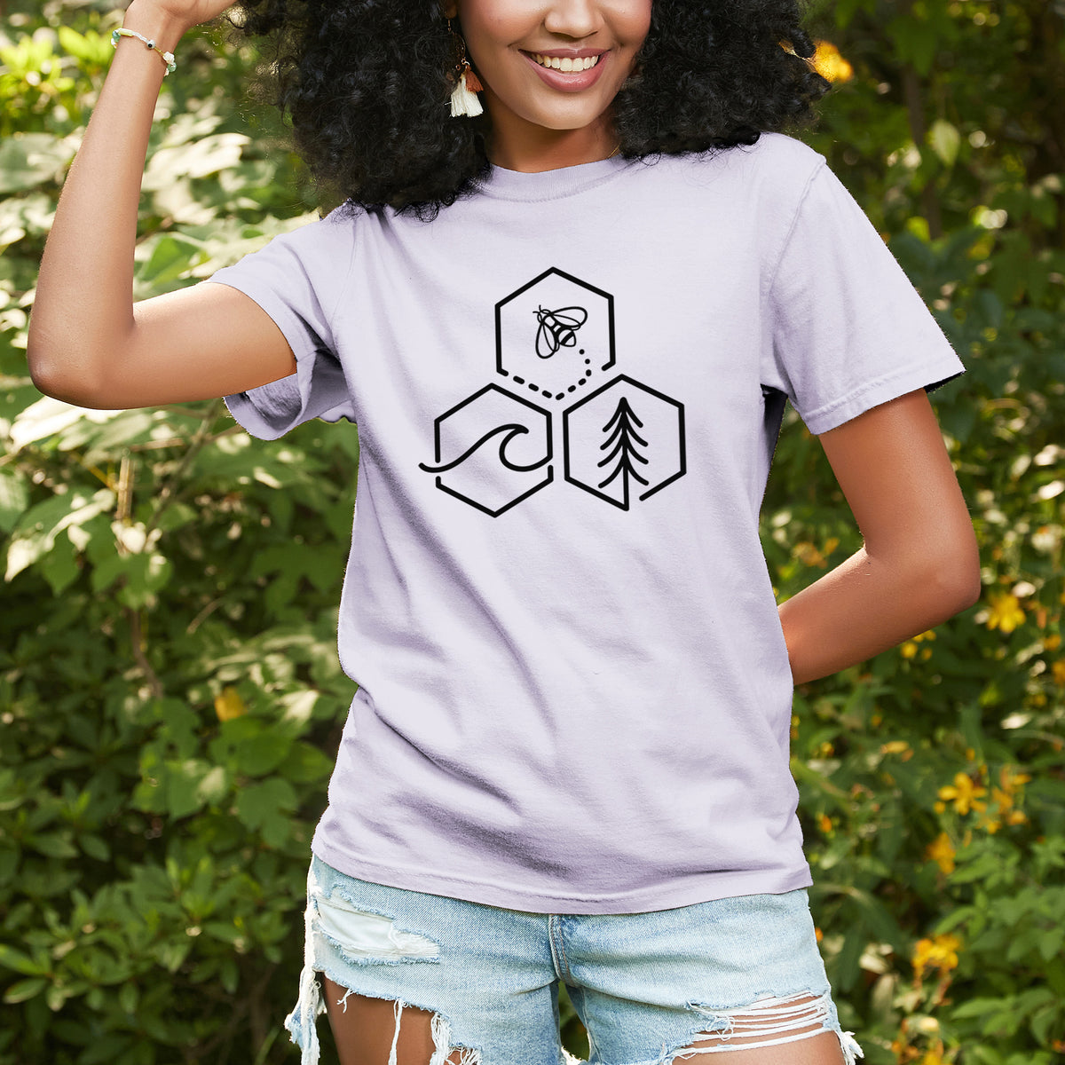 Woman wearing a Comfort Colors t-shirt printed with a black line art design of three hexagons holding a bee, an ocean wave and a pine tree, outdoors.