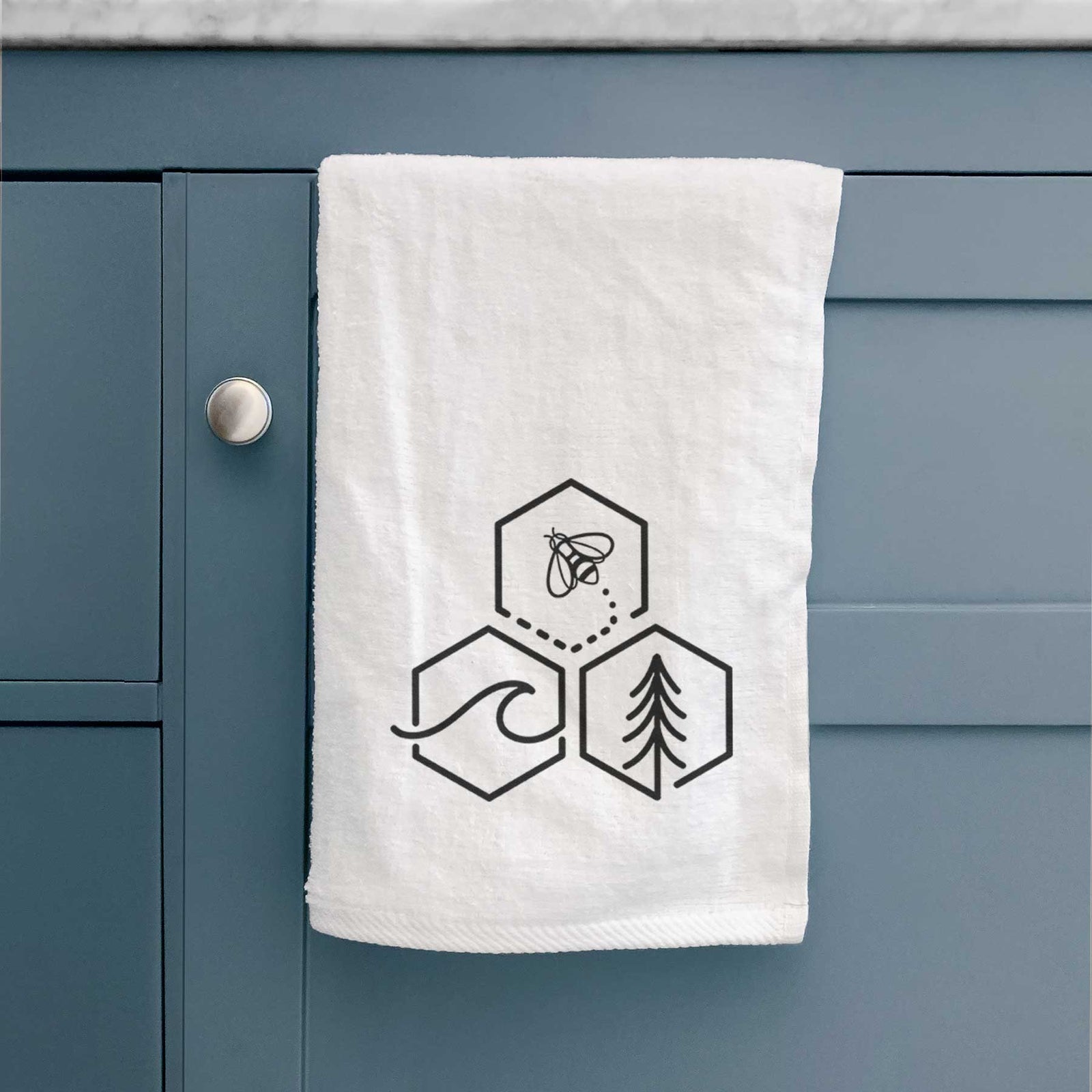 White hand towel printed with a black line art design of three hexagons holding a bee, an ocean wave and a pine tree.