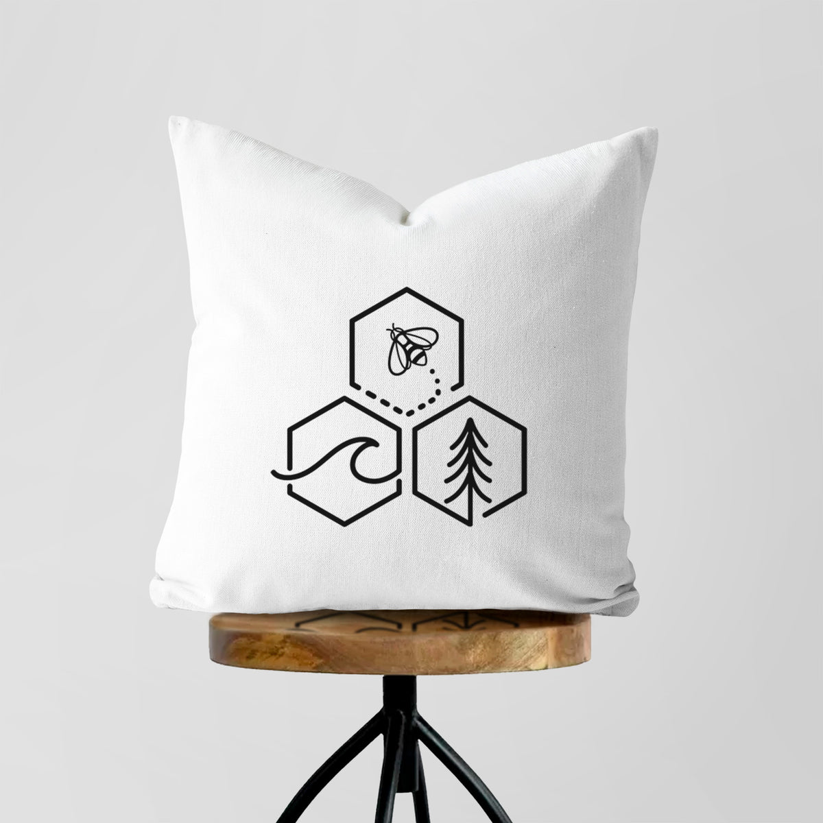 White pillow cover printed with a black line art design of three hexagons holding a bee, an ocean wave and a pine tree, on a wooden stool.