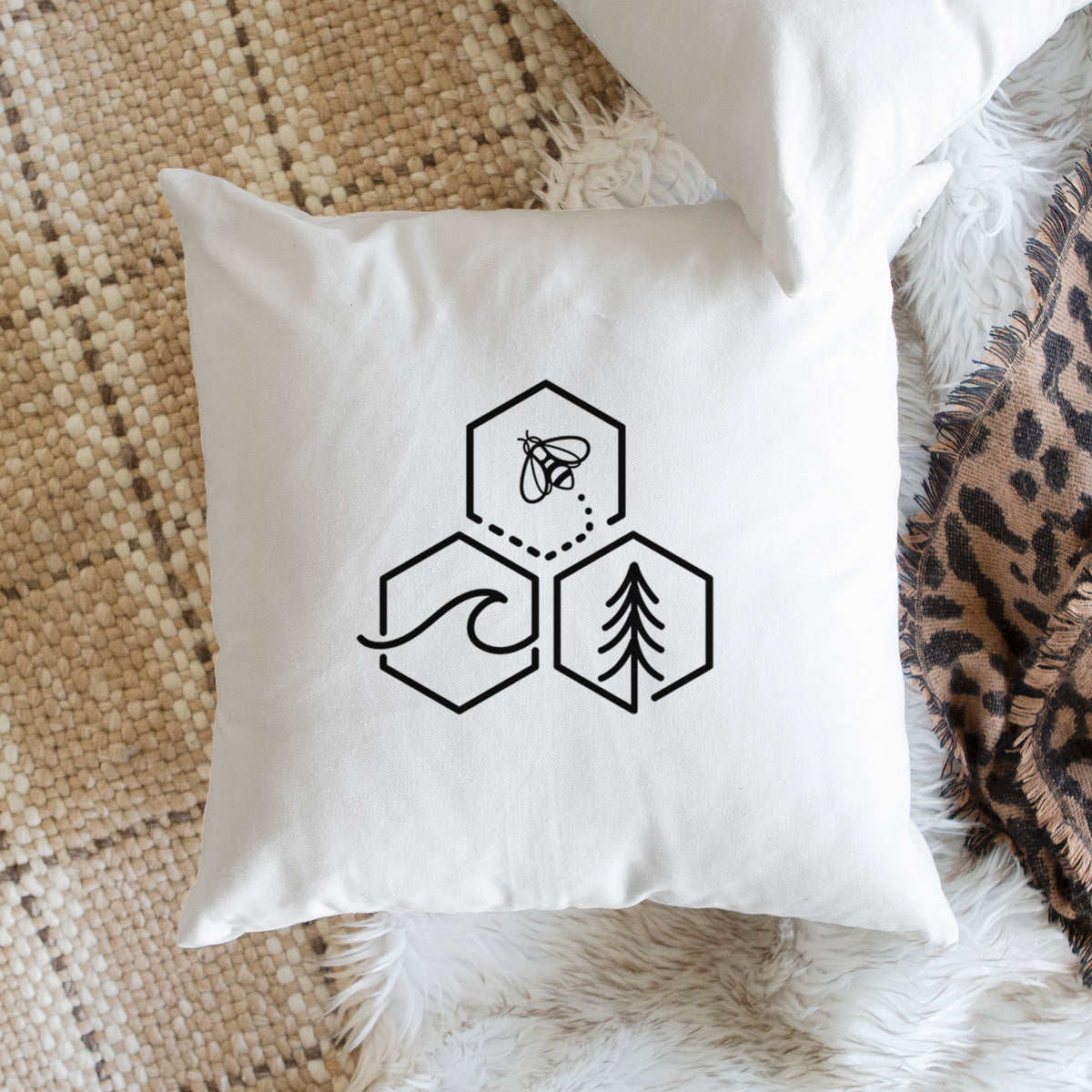 White pillow cover printed with a black line art design of three hexagons holding a bee, an ocean wave and a pine tree, on a woven chair.