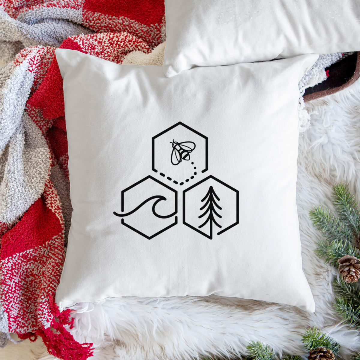 White pillow cover printed with a black line art design of three hexagons holding a bee, an ocean wave and a pine tree, on a woven chair.